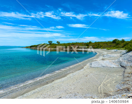 Sunny day at a beach with clear water and a shore lined with pebbles and rocks near green trees in the background Sunny day at a beach with clear water and a shore lined with pebbles and rocks near green trees in the background 140650984