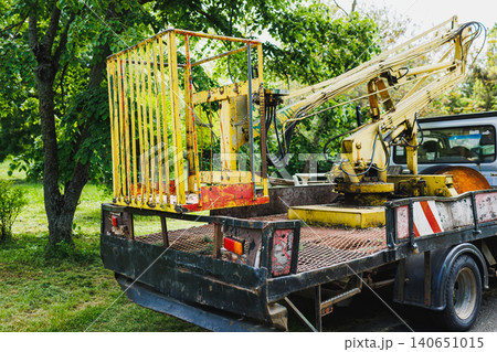 Park maintenance truck with elevated lift, rusted bucket secured on flatbed, folded crane boom, leafy backdrop, soft morning light, equipment staged for arborist repairs 140651015