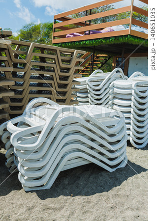 Wooden platform features neatly stacked sun loungers and gravel pathways. Sundrenched coastal space displays carefully arranged chairs and storage units prepared for visitors Wooden platform features neatly stacked sun loungers and gravel pathways. Sundrenched coastal space displays carefully arranged chairs and storage units prepared for visitors 140651035