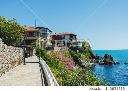 Sunny coastal walk. Bright seaside path with flowers. Radiant cliff trail overlooking calm waters. Sunshine illuminating historic houses along tranquil harbor scene Sunny coastal walk. Bright seaside path with flowers. Radiant cliff trail overlooking calm waters. Sunshine illuminating historic houses along tranquil harbor scene 140651216