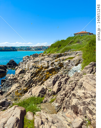 Rocky coastline with blue water and a house on the cliff near a sunny beach during midday 140651248