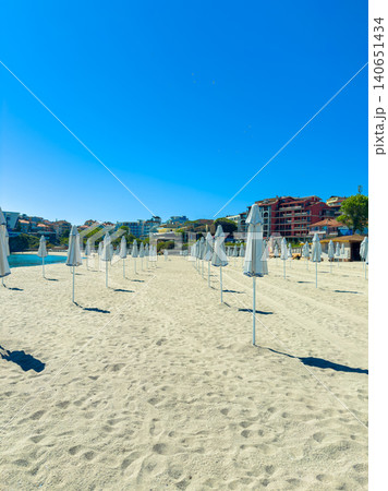 Sunshine day at the beach with many umbrellas lined up in the sand and clear blue sky in a coastal area near city buildings during daytime Sunshine day at the beach with many umbrellas lined up in the sand and clear blue sky in a coastal area near city buildings during daytime 140651434