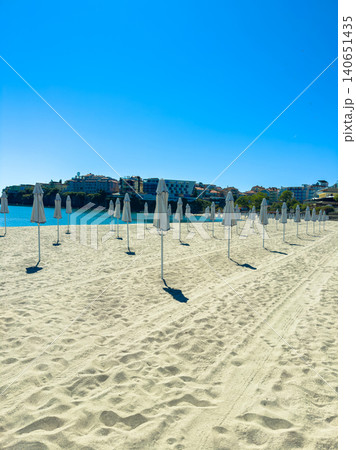 Empty beach with rows of umbrellas under bright blue sky near a coastal town during midday Empty beach with rows of umbrellas under bright blue sky near a coastal town during midday 140651435