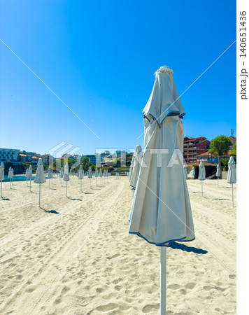 Bright beach scene with umbrellas arranged neatly in sand during daylight, showing clear sky and coastal buildings in the background 140651436