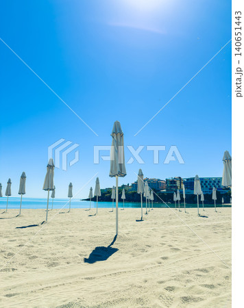 Empty beach with many umbrellas standing under a clear blue sky at midday near a coastal building Empty beach with many umbrellas standing under a clear blue sky at midday near a coastal building 140651443