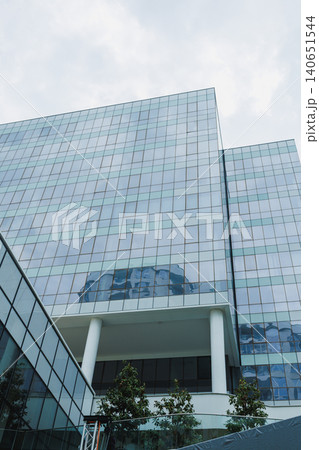Modern glass building stands tall against a cloudy sky in a busy city environment filled with trees and greenery Modern glass building stands tall against a cloudy sky in a busy city environment filled with trees and greenery 140651544