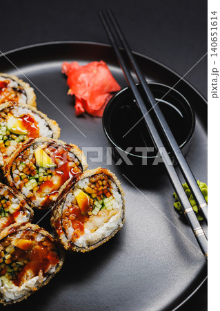 Enjoying sushi rolls served with soy sauce and garnishes on a black plate at a dining table during dinner time in a home setting Enjoying sushi rolls served with soy sauce and garnishes on a black plate at a dining table during dinner time in a home setting 140651614