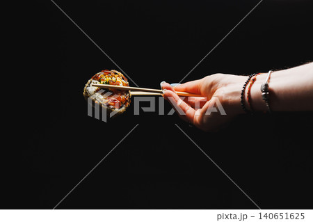 Hand holds sushi with chopsticks above dark surface while highlighting the intricate details of the roll in a simple indoor space during a meal time Hand holds sushi with chopsticks above dark surface while highlighting the intricate details of the roll in a simple indoor space during a meal time 140651625
