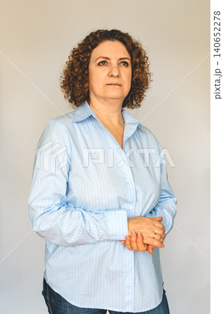 Thoughtful middle-aged woman in blue shirt standing with folded hands for portrait Thoughtful middle-aged woman in blue shirt standing with folded hands for portrait 140652278