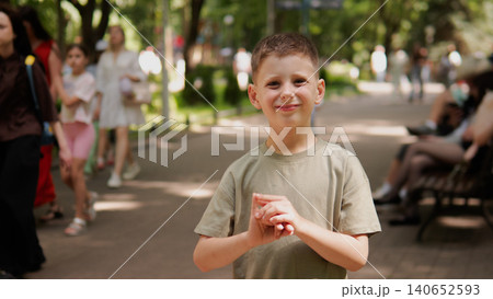 A young boy stands in a sunny park, smiling at the camera with hands gently clasped in front. Dappled shade, trees, and a blurred crowd create a lively outdoor setting A young boy stands in a sunny park, smiling at the camera with hands gently clasped in front. Dappled shade, trees, and a blurred crowd create a lively outdoor setting 140652593