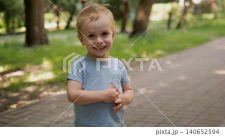 A young boy wearing a light blue T-shirt stands on a paved park path with trees and sunlight in the background, hands clasped softly, presenting a cheerful and relaxed outdoor A young boy wearing a light blue T-shirt stands on a paved park path with trees and sunlight in the background, hands clasped softly, presenting a cheerful and relaxed outdoor 140652594