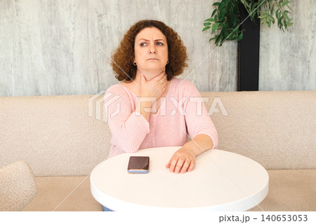 Middle-aged woman sitting at a table touching her throat with a worried expression and throat pain 140653053