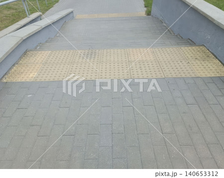 Top down aerial view of a paved sloping walkway with yellow tactile paving strips, handrails on both sides, and green grass bordering the path... 140653312