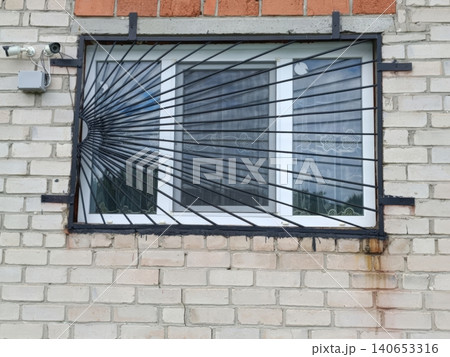 A white two-lens security camera mounted on a brick wall above a window with heavy black metal bars and grate. The building exterior shows signs of aging, creating a surveillance-focused urban scene A white two-lens security camera mounted on a brick wall above a window with heavy black metal bars and grate. The building exterior shows signs of aging, creating a surveillance-focused urban scene 140653316