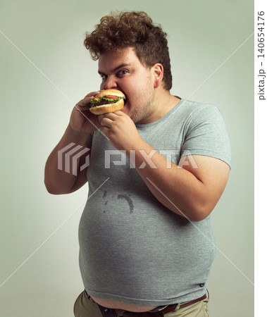 Obesity, weight gain and hamburger for man in studio with food for unhealthy eating, plus size and greed. Overweight person with meal in mouth and fat stomach for expression humor and comedy indoor 140654176