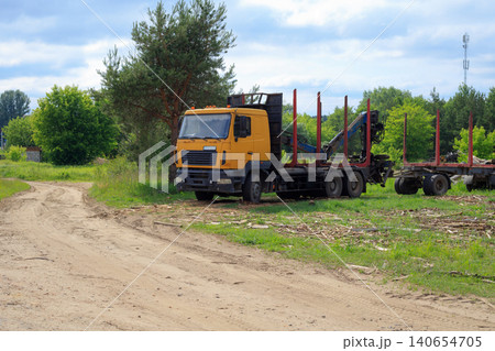 Yellow timber truck with crane arm parked near dirt road, empty log carrier semi trailer on green glade landscape Yellow timber truck with crane arm parked near dirt road, empty log carrier semi trailer on green glade landscape 140654705