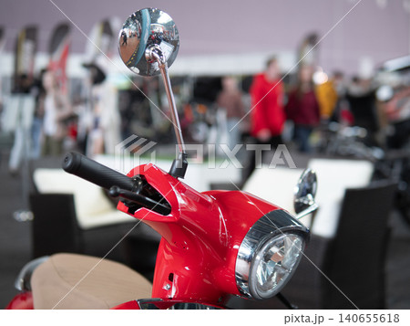 Red Scooter Front In Showroom, Glossy Chrome Mirror And Headlight In Sharp Focus, Blurred Shoppers And Display Booths In Background, Modern Lifestyle Composition With Retail Lighting Red Scooter Front In Showroom, Glossy Chrome Mirror And Headlight In Sharp Focus, Blurred Shoppers And Display Booths In Background, Modern Lifestyle Composition With Retail Lighting 140655618