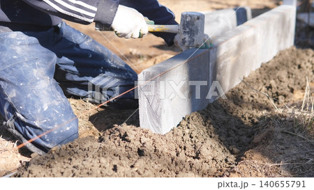 Mason Setting Curb Block Using Mallet And Stringline For Final Positioning, Tapping Joints Into Alignment, Closeup Of Hands, Gloves And Block Edges, Finishing Touches For Pathway Border 140655791