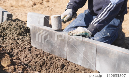 Worker Fitting Concrete Block Into Mortar Bed, Hands And Gloves Pressing Block For Secure Bond, Focus On Placement And Seam Alignment, Outdoor Site With Wheelbarrow And Soil, Careful Worker Fitting Concrete Block Into Mortar Bed, Hands And Gloves Pressing Block For Secure Bond, Focus On Placement And Seam Alignment, Outdoor Site With Wheelbarrow And Soil, Careful 140655807