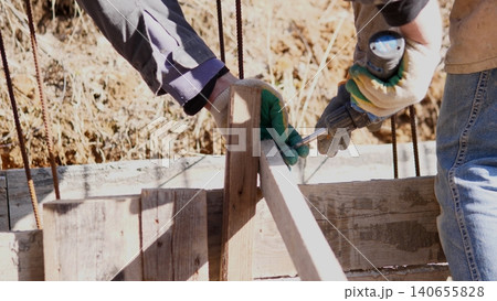 Hands Assembling Wooden Construction Formwork For Foundation, Workers Aligning Timber And Securing Rebar With Gloves And Drill At Muddy Excavation, Temporary Braces And Concrete Forms Hands Assembling Wooden Construction Formwork For Foundation, Workers Aligning Timber And Securing Rebar With Gloves And Drill At Muddy Excavation, Temporary Braces And Concrete Forms 140655828
