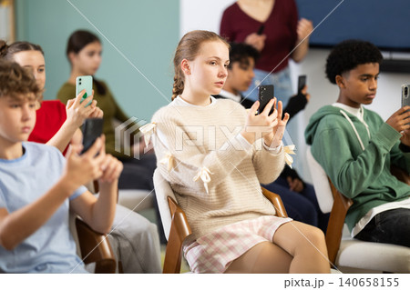 Group of teen listeners, girl take photos of presentation slide or scan QR code, teacher in background Group of teen listeners, girl take photos of presentation slide or scan QR code, teacher in background 140658155