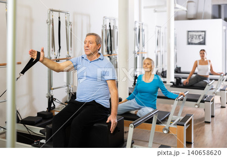 Serious old man in sportswear sitting and doing Pilates exercises for hands on reformer bed equipment in gym 140658620
