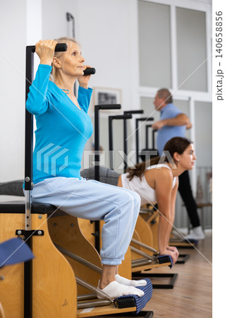 Focused old woman in sportswear training legs sitting and pressing down platform on Pilates Wanda chair equipment in rehabilitation center 140658856