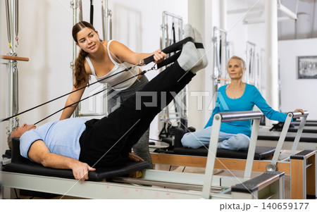 Positive young female instructor controlling movements of elderly man stretching on Pilates reformer equipment in studio 140659177