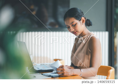 Concerned Asian businesswoman reviewing financial papers beside laptop in modern cafe office, showing budget problems, accounting pressure, data analysis, business risk, and difficult decisions. Concerned Asian businesswoman reviewing financial papers beside laptop in modern cafe office, showing budget problems, accounting pressure, data analysis, business risk, and difficult decisions. 140659315