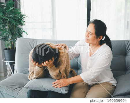 Caring Asian woman comforting a sad young woman on a sofa with a pillow, showing family support, empathy, stress relief, reassurance, mental wellness, home care, and emotional healing. 140659494