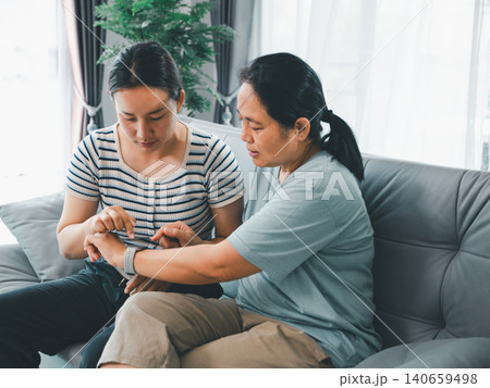 Asian women sitting on a home sofa checking a smartwatch together, showing family wellness, technology, health tracking, caregiving, digital support, lifestyle, and daily routine care. Asian women sitting on a home sofa checking a smartwatch together, showing family wellness, technology, health tracking, caregiving, digital support, lifestyle, and daily routine care. 140659498
