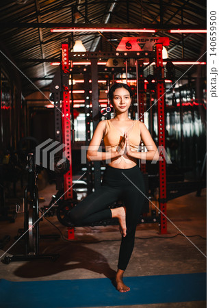 Athletic Asian woman standing in tree pose on a blue yoga mat inside a dark gym, prayer hands at chest, showing balance, focus, strength, mindful fitness, stability, and wellness discipline. Athletic Asian woman standing in tree pose on a blue yoga mat inside a dark gym, prayer hands at chest, showing balance, focus, strength, mindful fitness, stability, and wellness discipline. 140659500