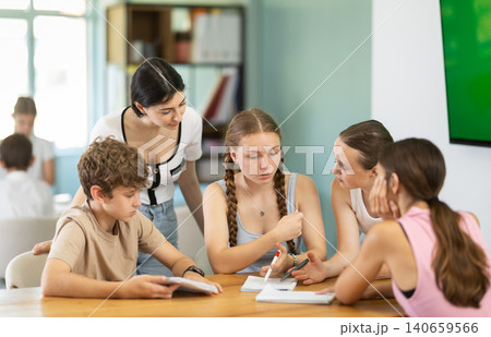Teenage students discussing assignments around table monitored by teacher Teenage students discussing assignments around table monitored by teacher 140659566