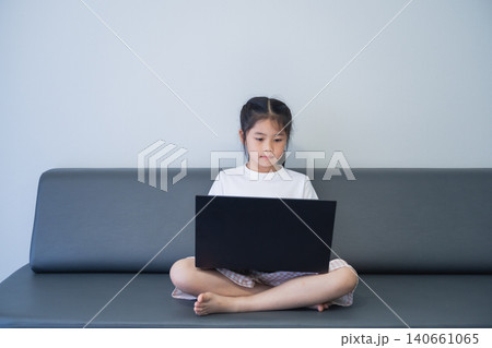 Young girl sitting on a modern gray sofa cross-legged, using a black laptop with a plain white wall background, demonstrating focus and digital technology use 140661065