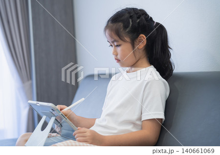 Young Girl Using Tablet Device Sitting on Sofa In Living Room Playing Educational Game or Watching Video With Pen in Hand Indoors 140661069