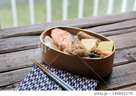 窓辺に置いた曲げわっぱに入れた焼き鮭の和食弁当と矢絣柄の包み布 140663170