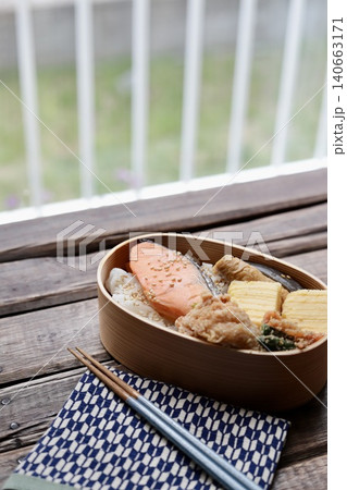 窓辺に置いた曲げわっぱに入れた焼き鮭の和食弁当と矢絣柄の包み布 140663171