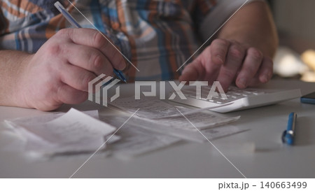 A person is sitting at a table, writing notes with a pen while using a calculator. Receipts are laid out on the table showing the busy work environment in an office setting. A person is sitting at a table, writing notes with a pen while using a calculator. Receipts are laid out on the table showing the busy work environment in an office setting. 140663499
