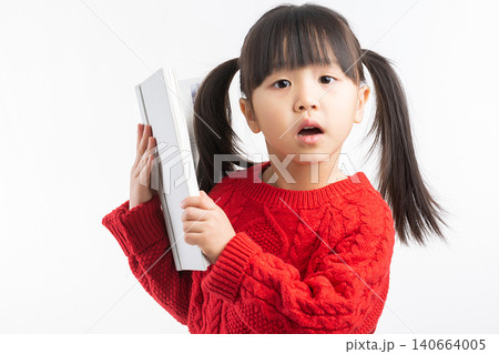 A surprised little Asian girl holding a large white book under her arm with her mouth open in astonishment. A surprised little Asian girl holding a large white book under her arm with her mouth open in astonishment. 140664005