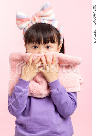 A studio shot of a young Asian girl with long dark hair and a striped headband, wearing a purple sweatshirt, playfully holding a pink hand towel in front of her face, partially obscuring it, against a A studio shot of a young Asian girl with long dark hair and a striped headband, wearing a purple sweatshirt, playfully holding a pink hand towel in front of her face, partially obscuring it, against a 140664289