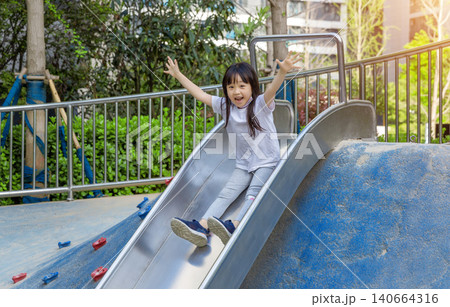 a cheerful girl sliding down a steel slide in the playground with open arms. a cheerful girl sliding down a steel slide in the playground with open arms. 140664316