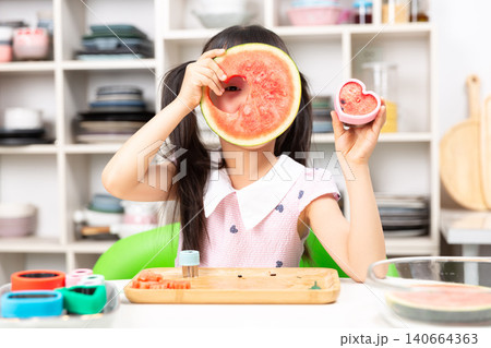 Playful young Asian girl covering one eye with watermelon slice while holding heart shaped toy in other hand Playful young Asian girl covering one eye with watermelon slice while holding heart shaped toy in other hand 140664363