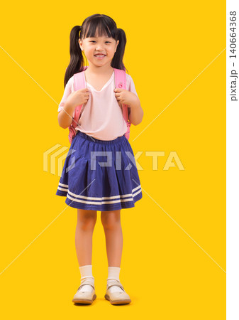 A cute Asian girl with a schoolbag on her back, a full-body portrait, stands in front of a yellow background in a photo studio. 140664368