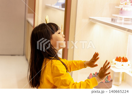 Curious little girl looks through a glass display case at beautiful multi-layered celebration cakes. Curious little girl looks through a glass display case at beautiful multi-layered celebration cakes. 140665224