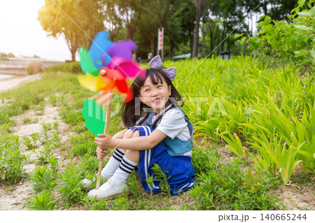 Happy Young East Asian Girl with Purple Bow Sitting in Sunny Green Meadow Holding Colorful Rainbow Pinwheel with Joyful Smile and Bright Summer Vibes. Happy Young East Asian Girl with Purple Bow Sitting in Sunny Green Meadow Holding Colorful Rainbow Pinwheel with Joyful Smile and Bright Summer Vibes. 140665244