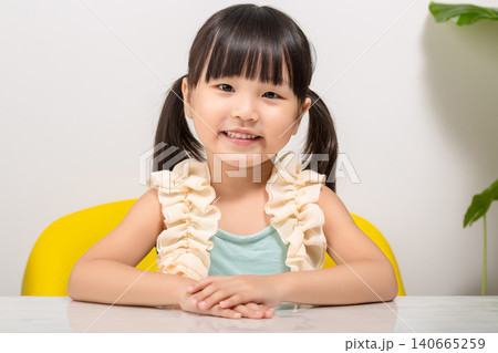 A happy and adorable Asian girl sits at a table in a bright room, smiling at the camera in a portrait photo. A happy and adorable Asian girl sits at a table in a bright room, smiling at the camera in a portrait photo. 140665259