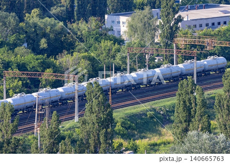 Freight train carrying cargo moves through a green landscape with trees and buildings in the background Freight train carrying cargo moves through a green landscape with trees and buildings in the background 140665763