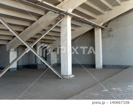 Empty industrial space underneath concrete stadium grandstands, featuring massive support pillars, steel cross-bracing beams, and a paved brick ground. 140667106