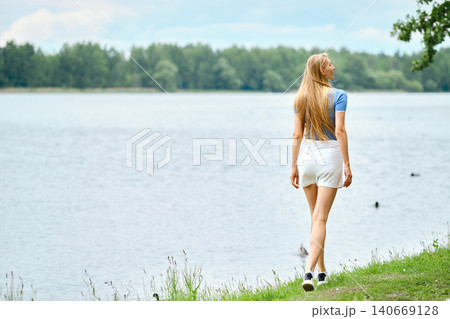 Woman walks along lake edge in summer near trees and water under cloudy sky Woman walks along lake edge in summer near trees and water under cloudy sky 140669128