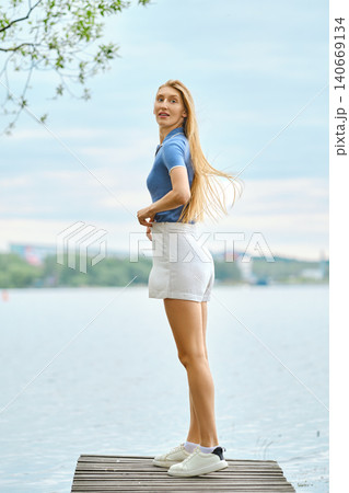 Woman in blue shirt and white shorts looks back standing on wooden pier by lake Woman in blue shirt and white shorts looks back standing on wooden pier by lake 140669134